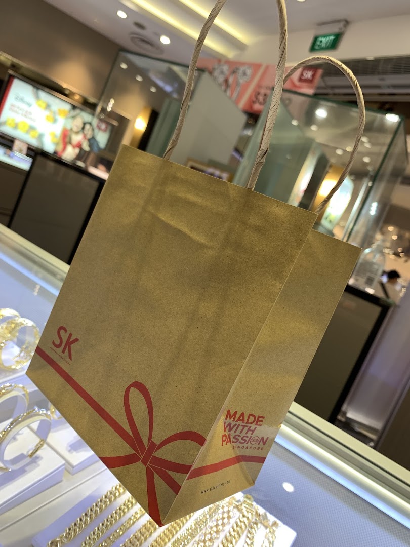 An SK Jewellery shopping bag with a red ribbon design, set against sparkling jewelry displays.