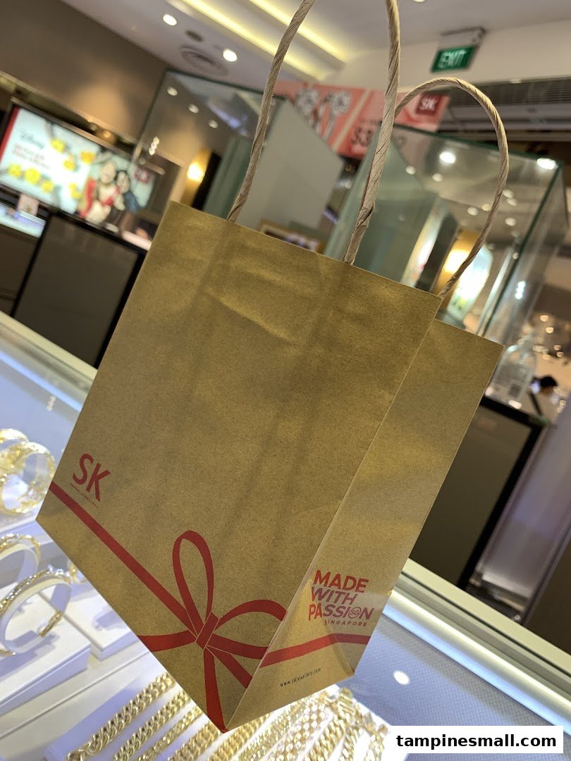 An SK Jewellery shopping bag with a red ribbon design, set against sparkling jewelry displays.