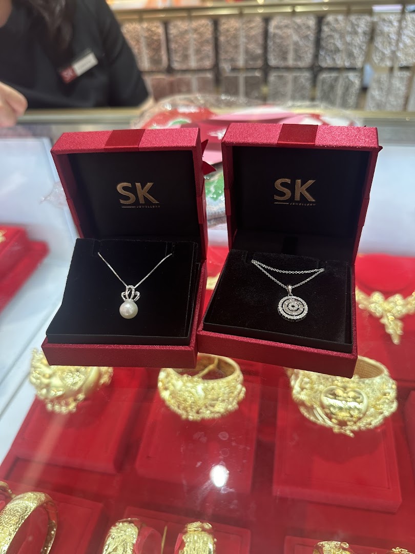 Two SK Jewellery necklace boxes displaying a pearl and a diamond pendant, on a red counter with gold jewelry.