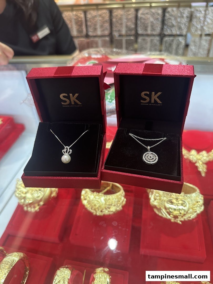 Two SK Jewellery necklace boxes displaying a pearl and a diamond pendant, on a red counter with gold jewelry.