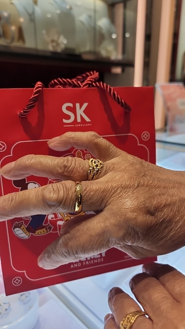 A hand adorned with gold rings holds an SK Jewellery shopping bag inside the store.