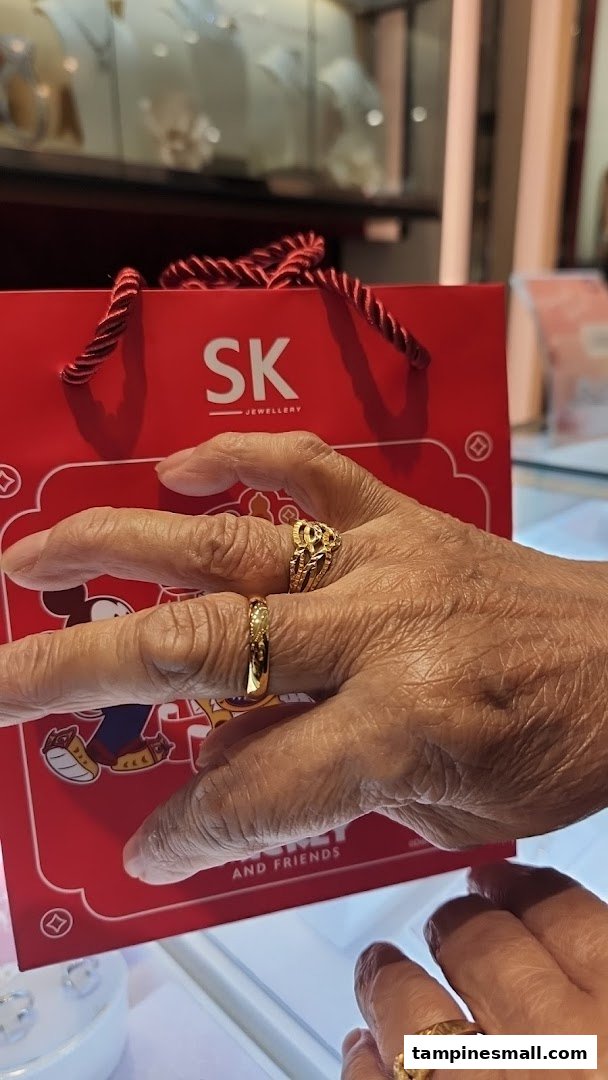 A hand adorned with gold rings holds an SK Jewellery shopping bag inside the store.