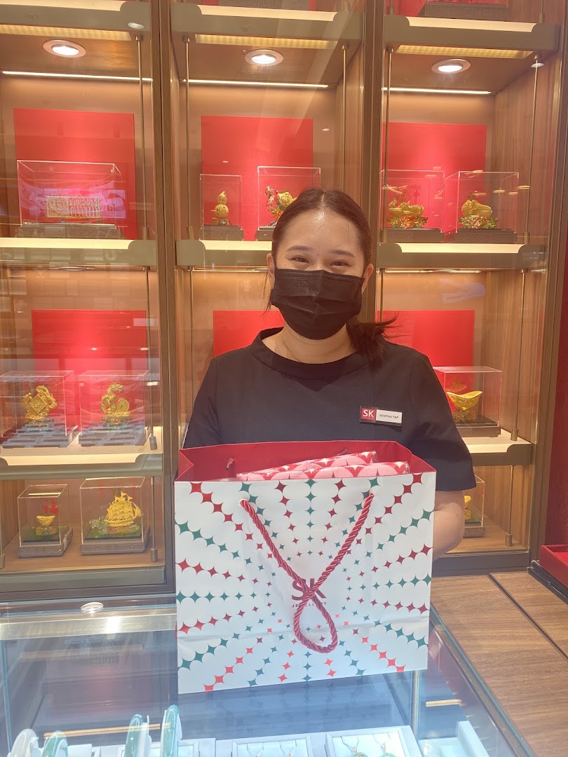 A smiling SK Jewellery staff member holds a branded shopping bag in front of gold displays.