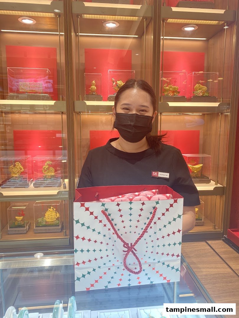A smiling SK Jewellery staff member holds a branded shopping bag in front of gold displays.