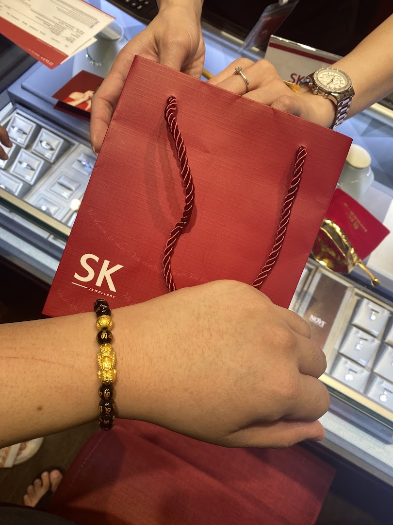 A customer tries on a gold bracelet while receiving a red SK Jewellery shopping bag in-store.