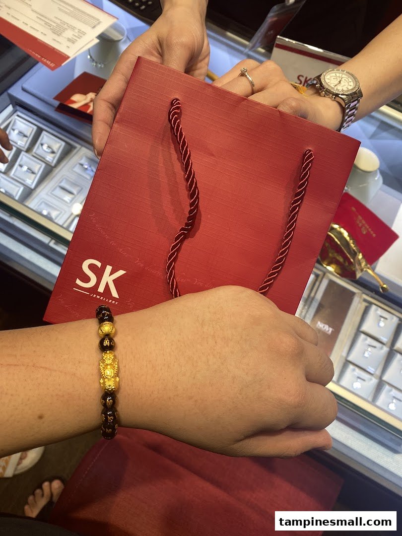 A customer tries on a gold bracelet while receiving a red SK Jewellery shopping bag in-store.