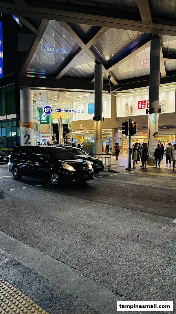Evening view of Tampines Mall entrance, featuring StarHub and Uniqlo signs, with cars and pedestrians.