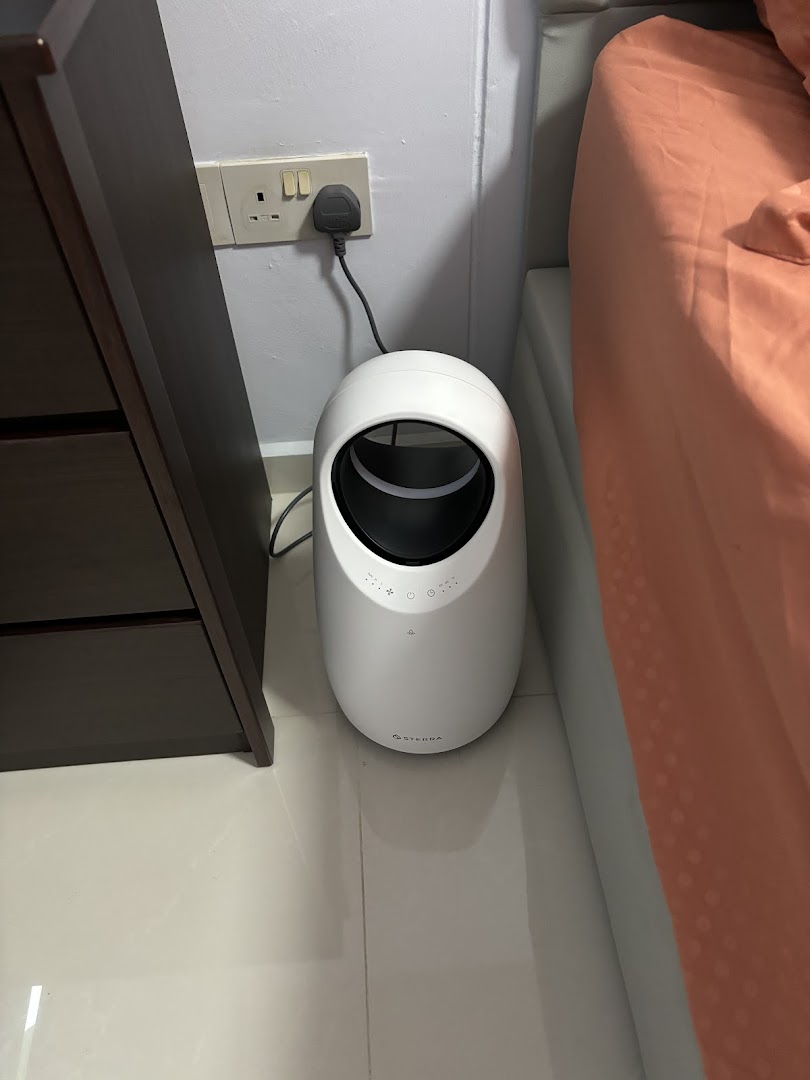A white Sterra product, likely an air purifier, plugged in next to a bed.