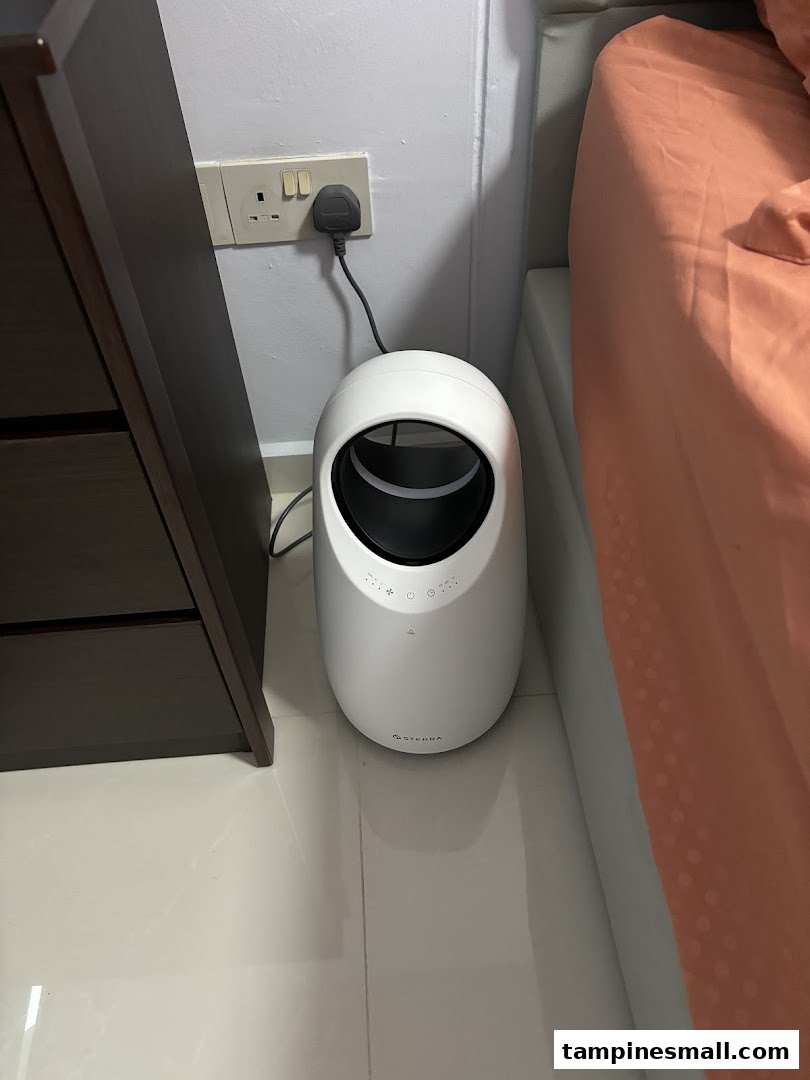 A white Sterra product, likely an air purifier, plugged in next to a bed.