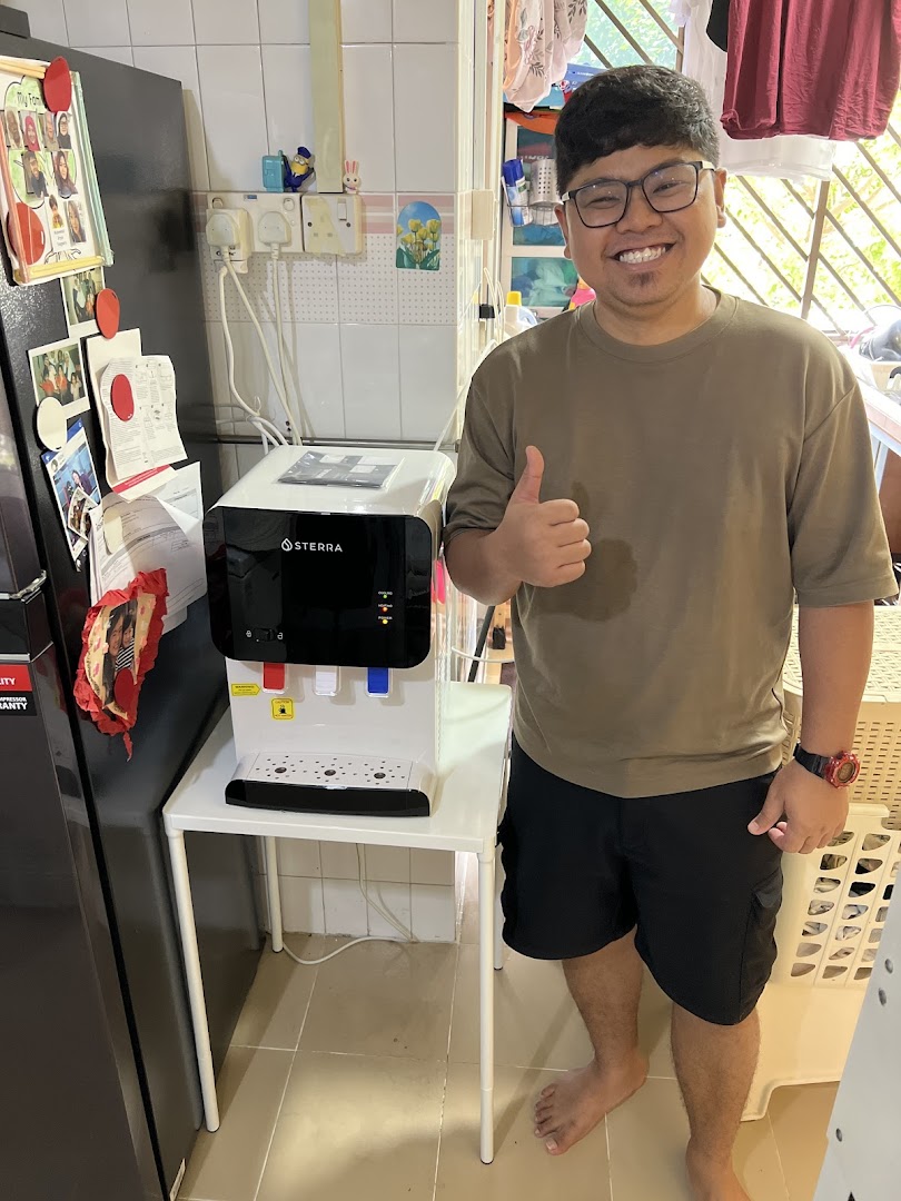 A smiling man gives a thumbs-up next to a Sterra water dispenser in a home setting.