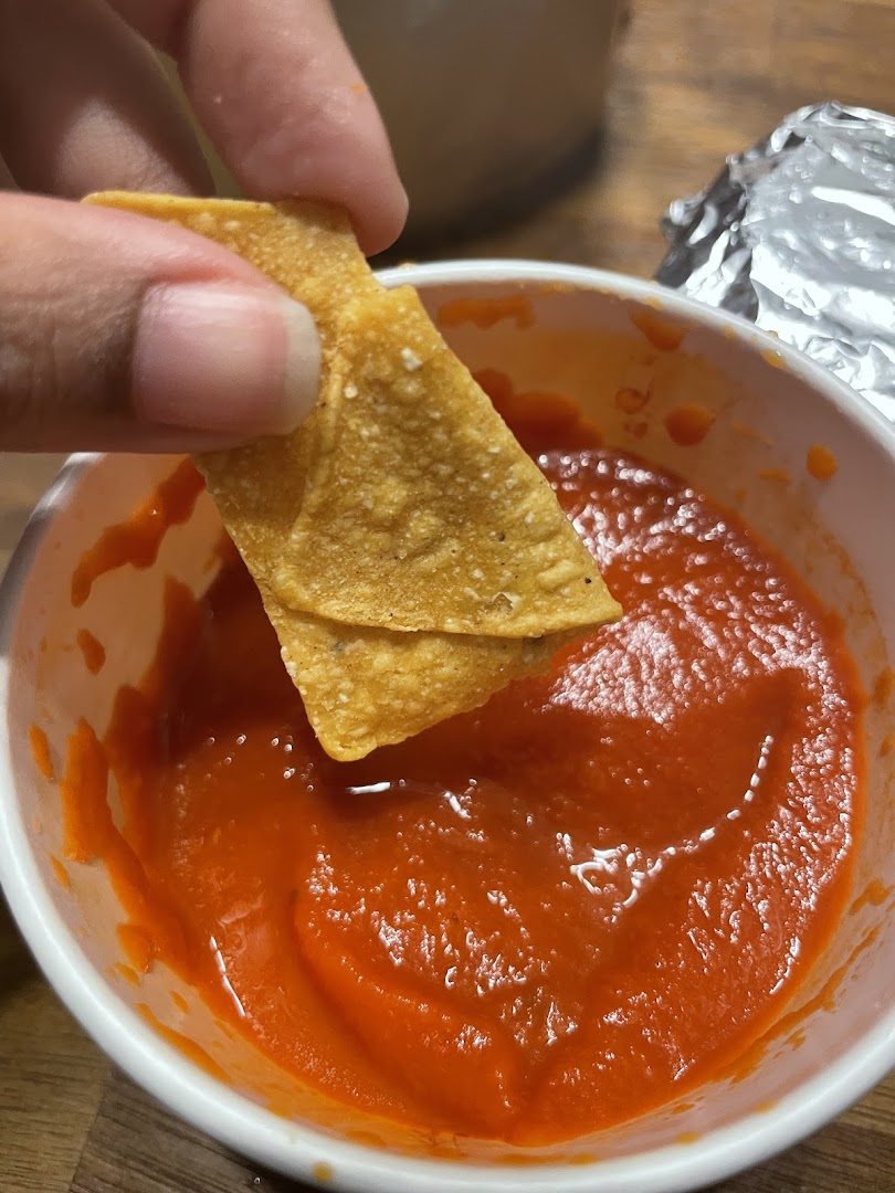 A hand dips a crispy tortilla chip into a vibrant red salsa in a white bowl.