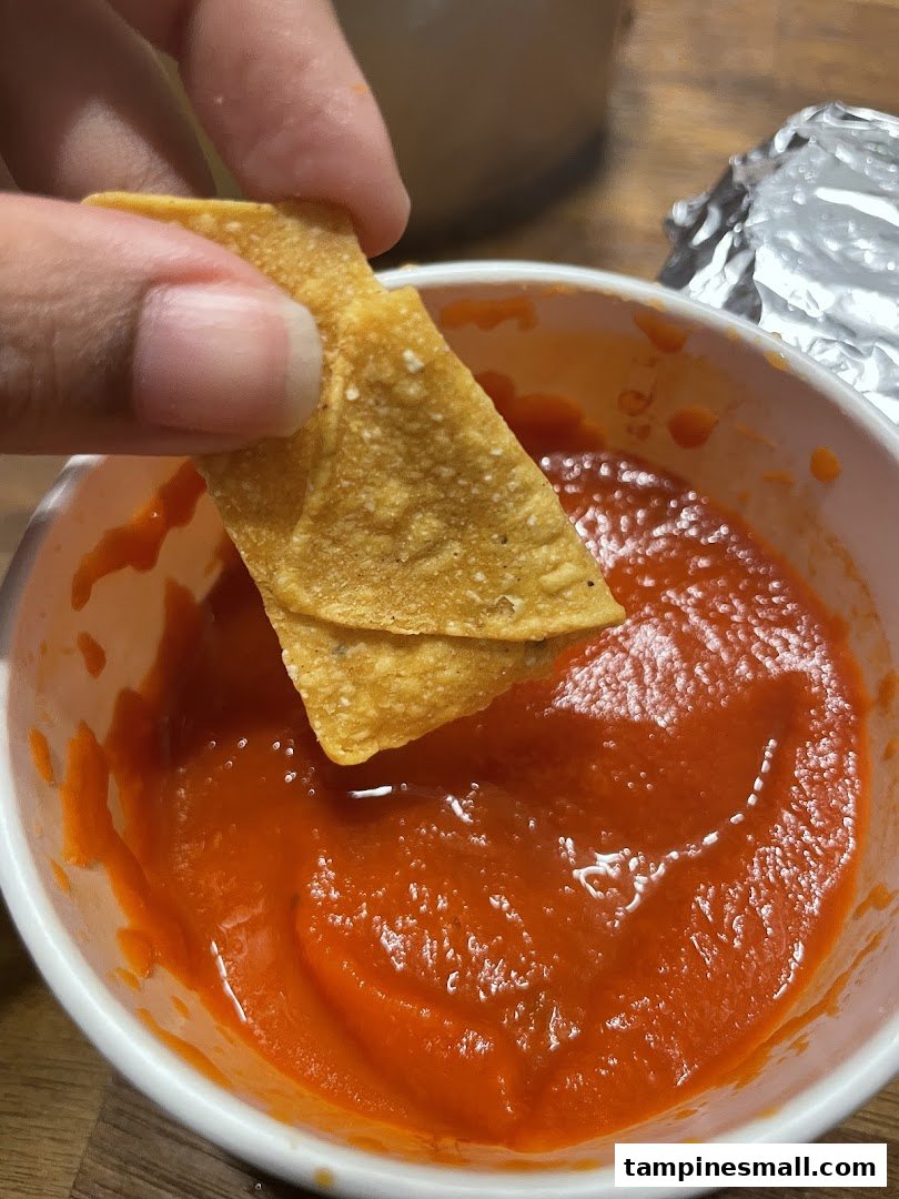 A hand dips a crispy tortilla chip into a vibrant red salsa in a white bowl.