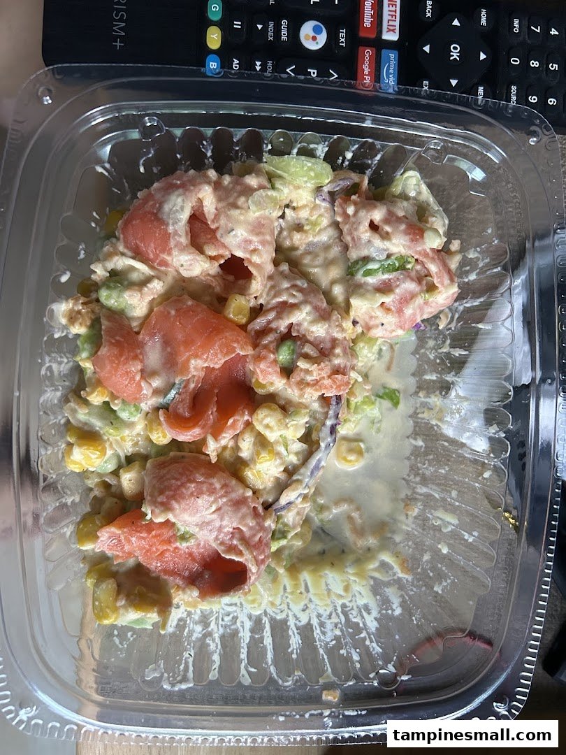 A close-up of a fresh salad or bowl with salmon, corn, and a creamy dressing in a clear plastic container.