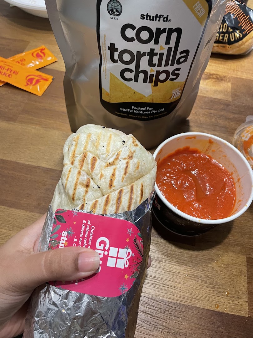 A hand holds a Stuffd wrap next to a cup of sauce and a bag of corn tortilla chips.