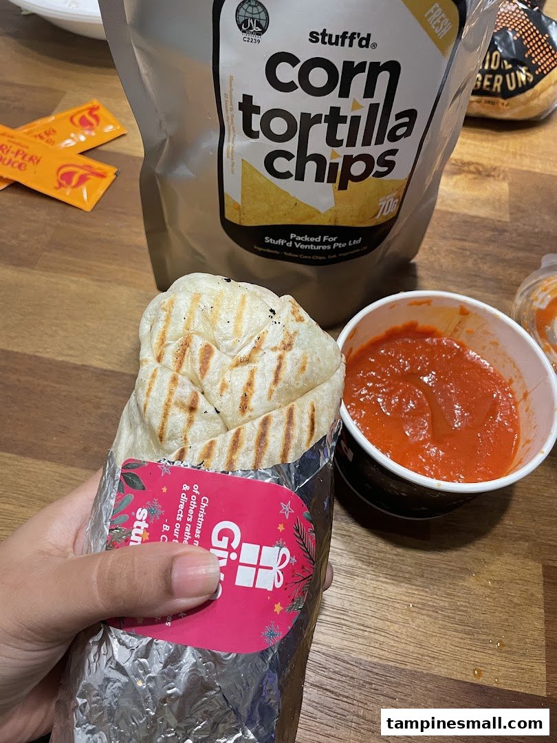 A hand holds a Stuffd wrap next to a cup of sauce and a bag of corn tortilla chips.