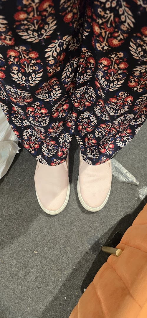 A person wearing patterned pants and light pink slip-on shoes stands on a grey carpet.