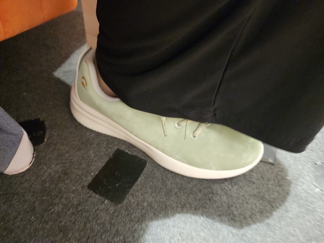 A close-up of a person's foot wearing a light green Sunnystep shoe on a dark carpeted floor.