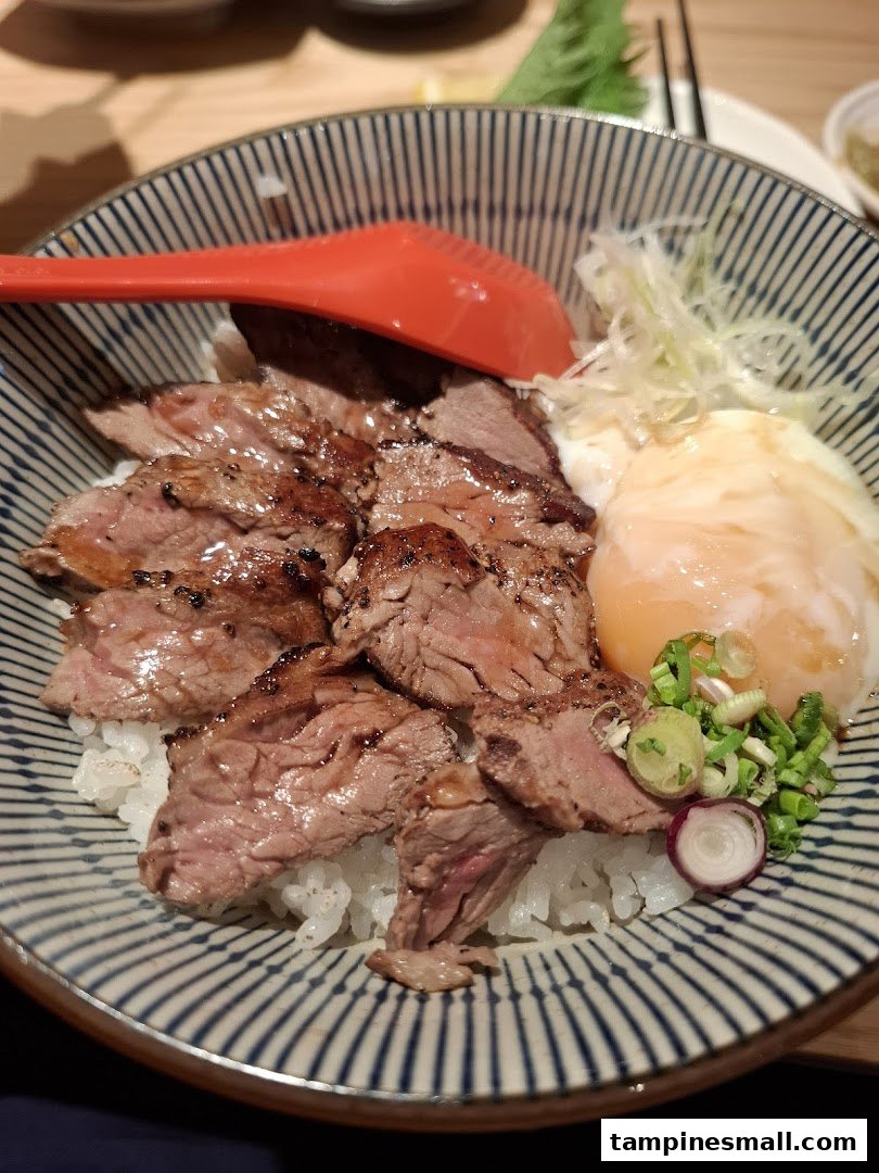 A delicious bowl of grilled beef slices with rice, a poached egg, and fresh garnishes.