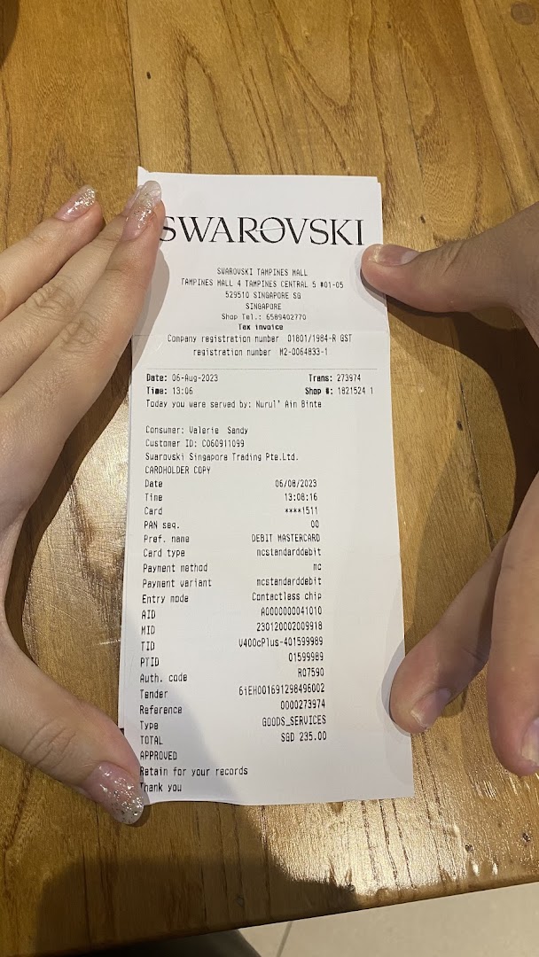 A pair of hands holding a detailed Swarovski retail receipt from Tampines Mall.