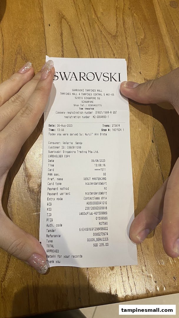 A pair of hands holding a detailed Swarovski retail receipt from Tampines Mall.