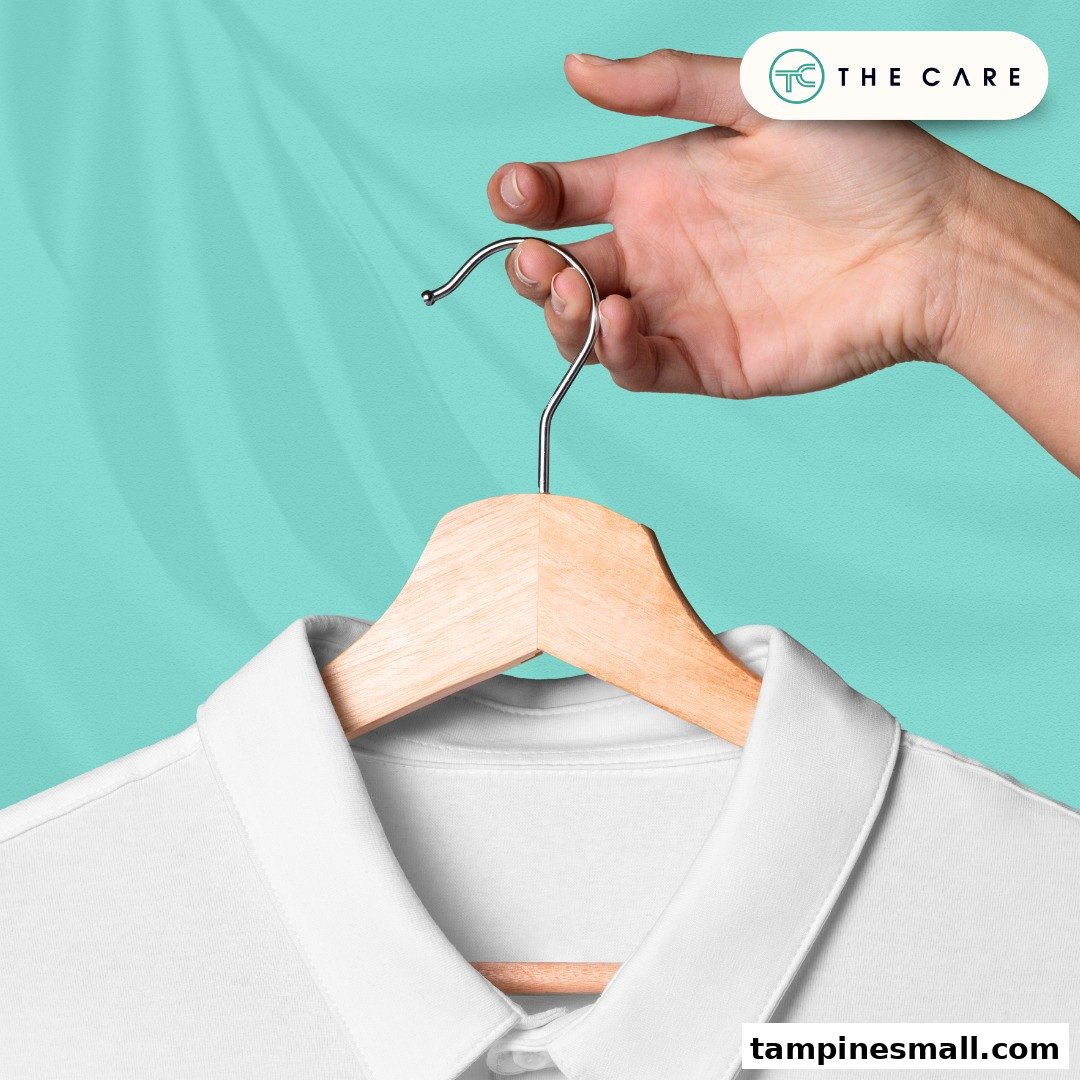 A hand holds a white shirt on a wooden hanger against a teal background, featuring the shop's logo.