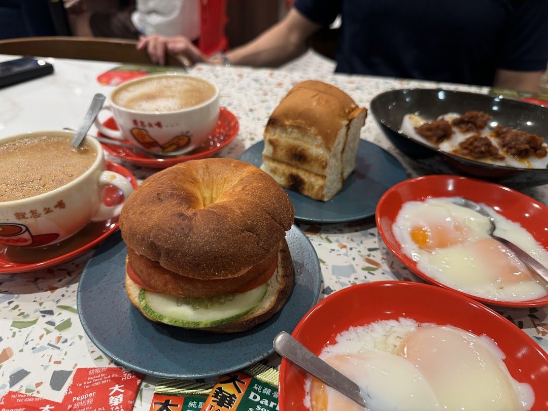 A table showcasing a delicious Hainanese breakfast spread with coffee, toast, eggs, and a bagel sandwich.
