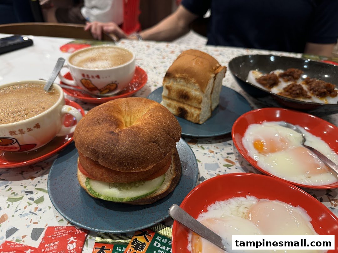 A table showcasing a delicious Hainanese breakfast spread with coffee, toast, eggs, and a bagel sandwich.