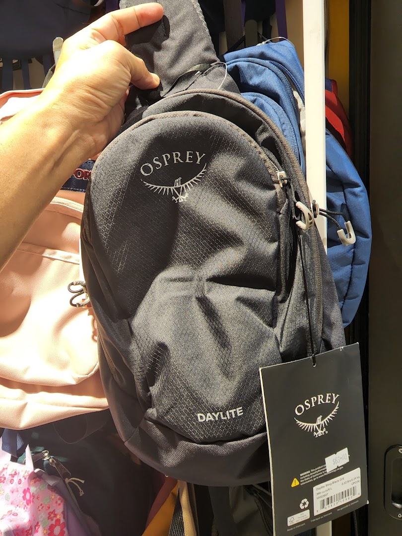 A hand holds a black Osprey Daylite sling bag with its price tag visible in a retail setting.