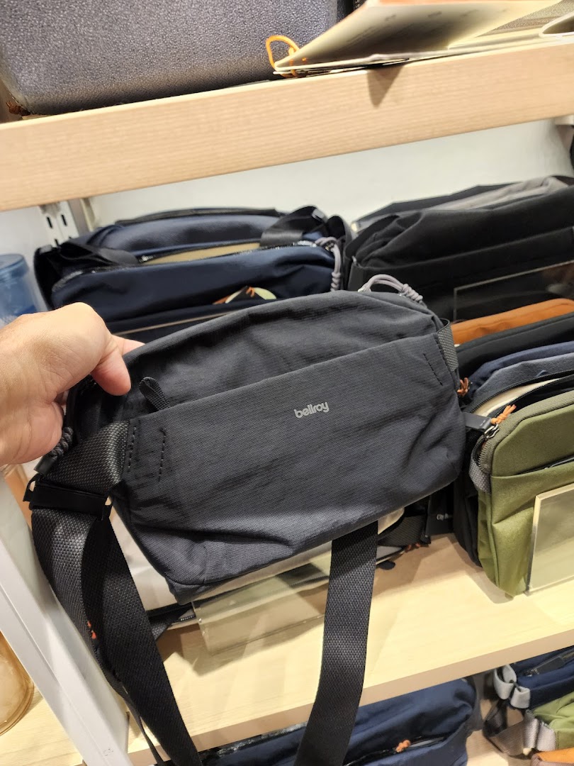 A hand holds a black Bellroy sling bag, showcasing products on shelves in a retail store.
