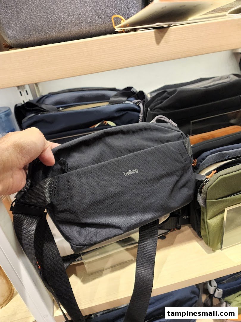 A hand holds a black Bellroy sling bag, showcasing products on shelves in a retail store.