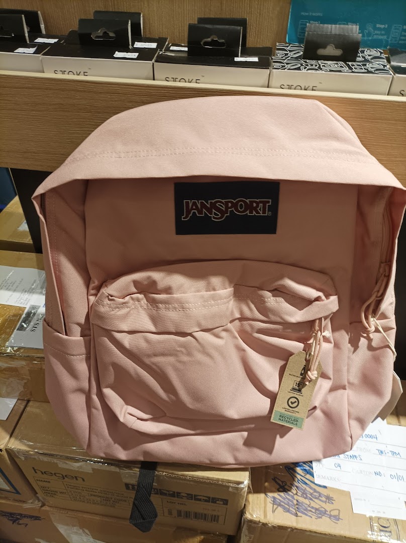 A light pink JanSport backpack with a 'Recycled Materials' tag, displayed in a retail setting.