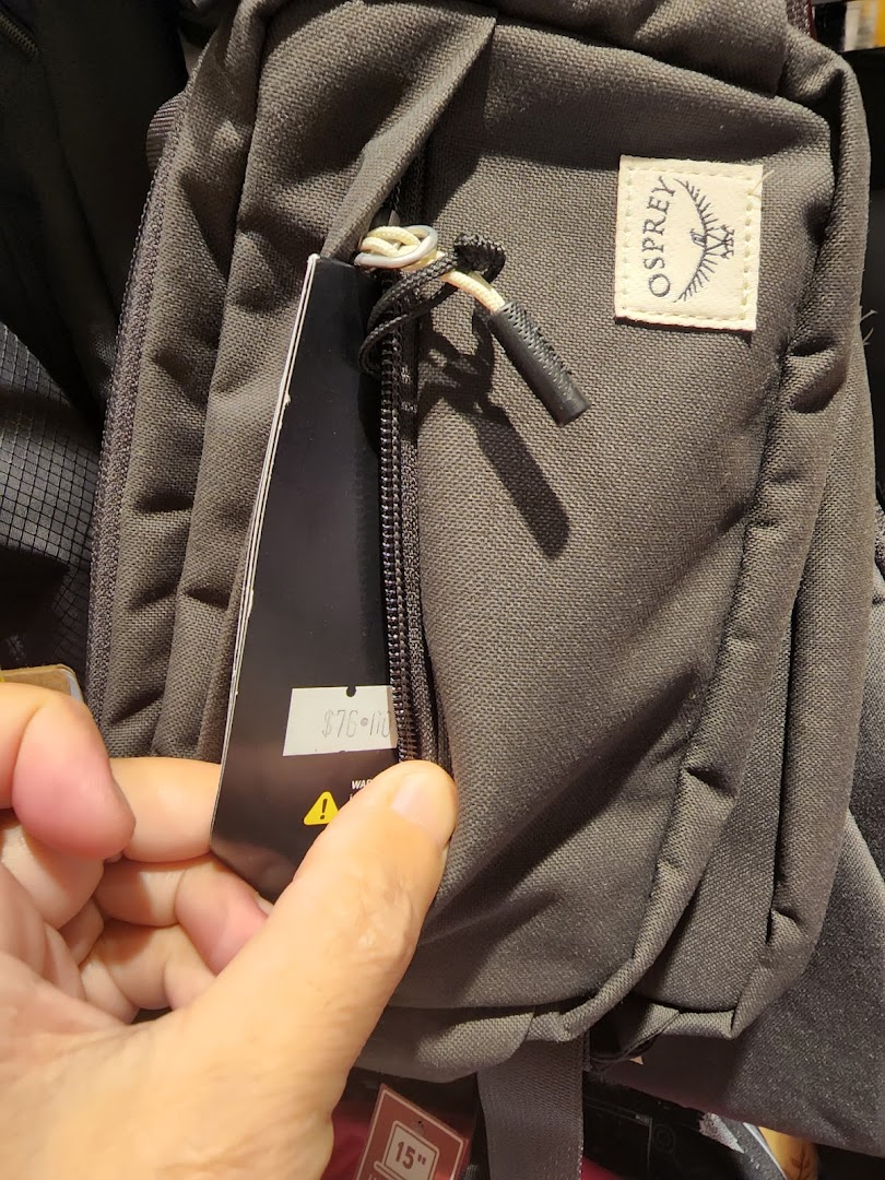 A hand holds a price tag on a dark grey Osprey bag, showcasing its features and price.