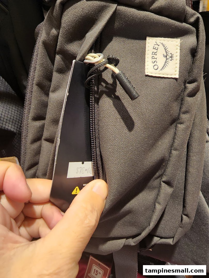 A hand holds a price tag on a dark grey Osprey bag, showcasing its features and price.