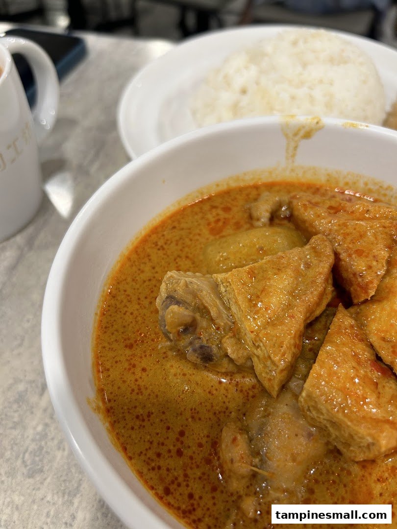 A close-up of a rich curry dish with chicken and tofu, served with rice and a beverage.