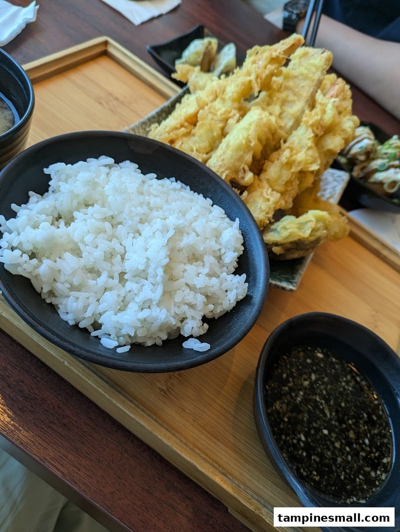 A close-up of a Japanese meal featuring crispy tempura, white rice, and a dipping sauce on a wooden tray.