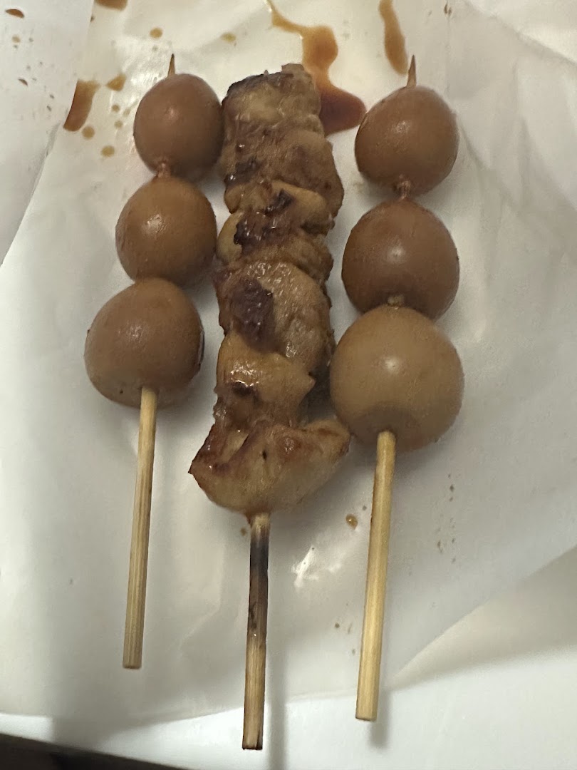 Close-up of three skewers, one with grilled chicken and two with quail eggs, served on white paper.
