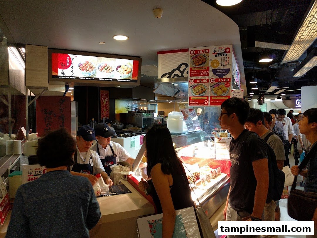 Customers queueing at the counter of TORI-Q, a Japanese food stall, with menu boards visible.