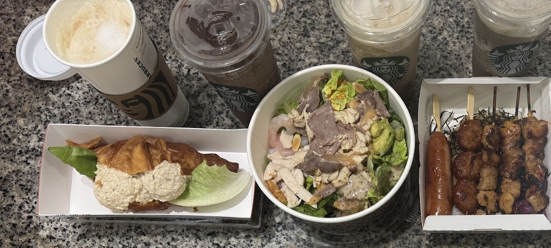 A diverse spread of takeout food and drinks, including skewers, a salad, a croissant sandwich, and Starbucks beverages.