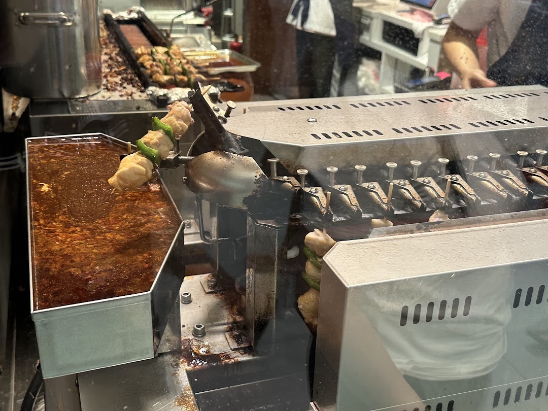 Skewers of food, possibly chicken or fish paste, cooking on an automated grill with a sauce bath.