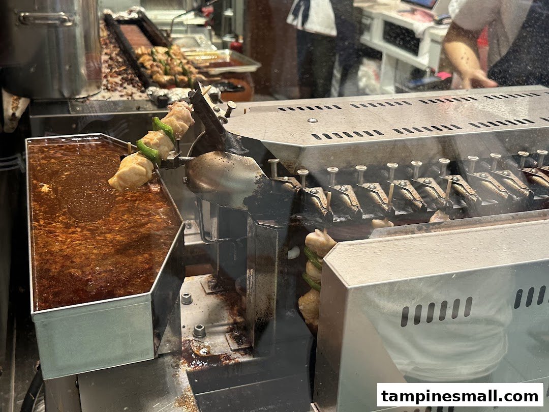 Skewers of food, possibly chicken or fish paste, cooking on an automated grill with a sauce bath.