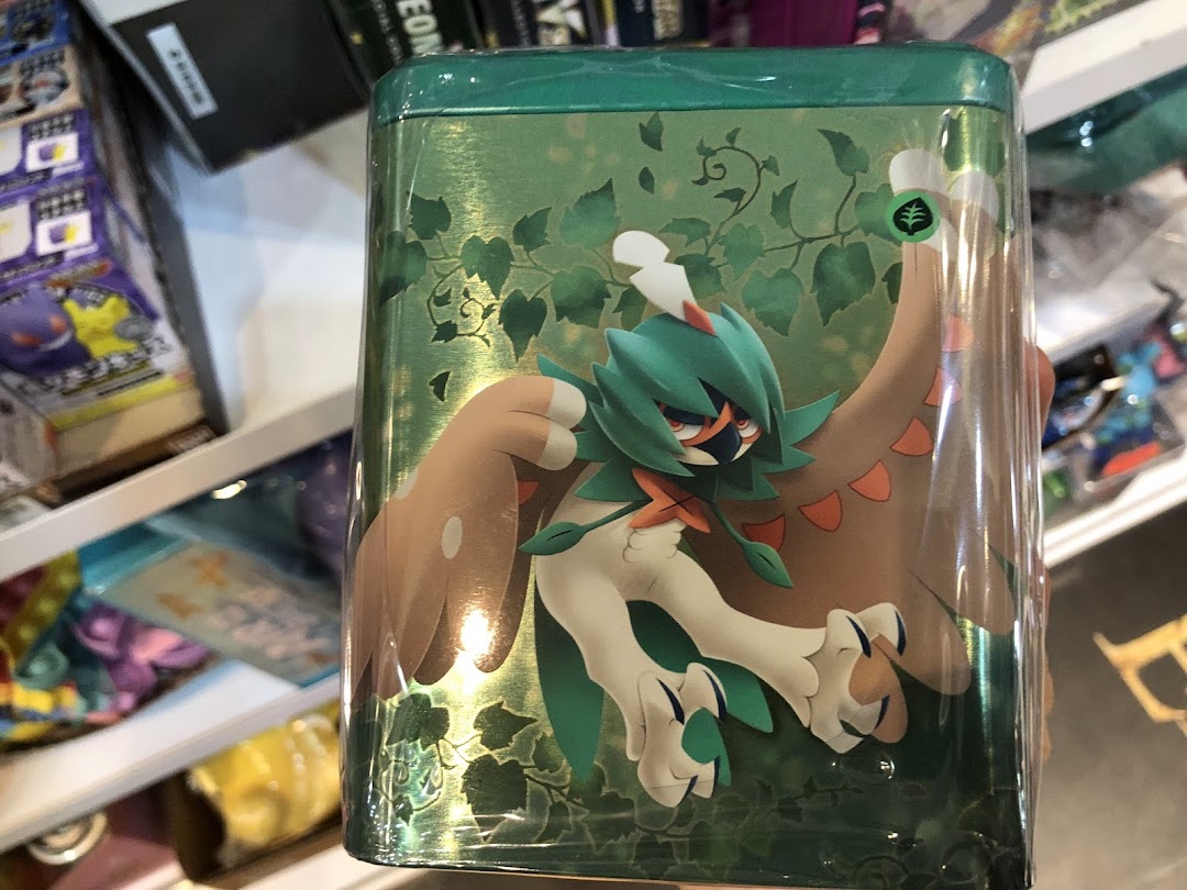 Close-up of a green Pokémon-themed tin featuring Decidueye, displayed in a toy or game shop.