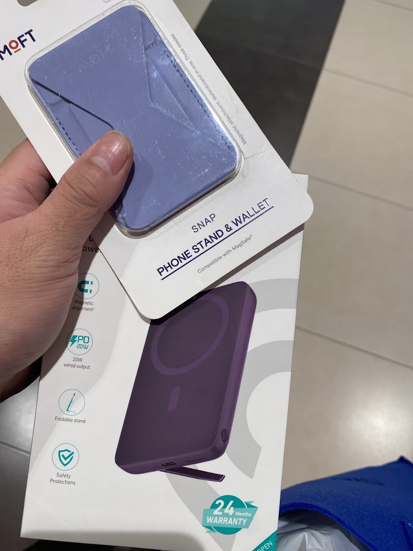 A hand holds two packaged tech accessories: a purple phone stand/wallet and a power bank.