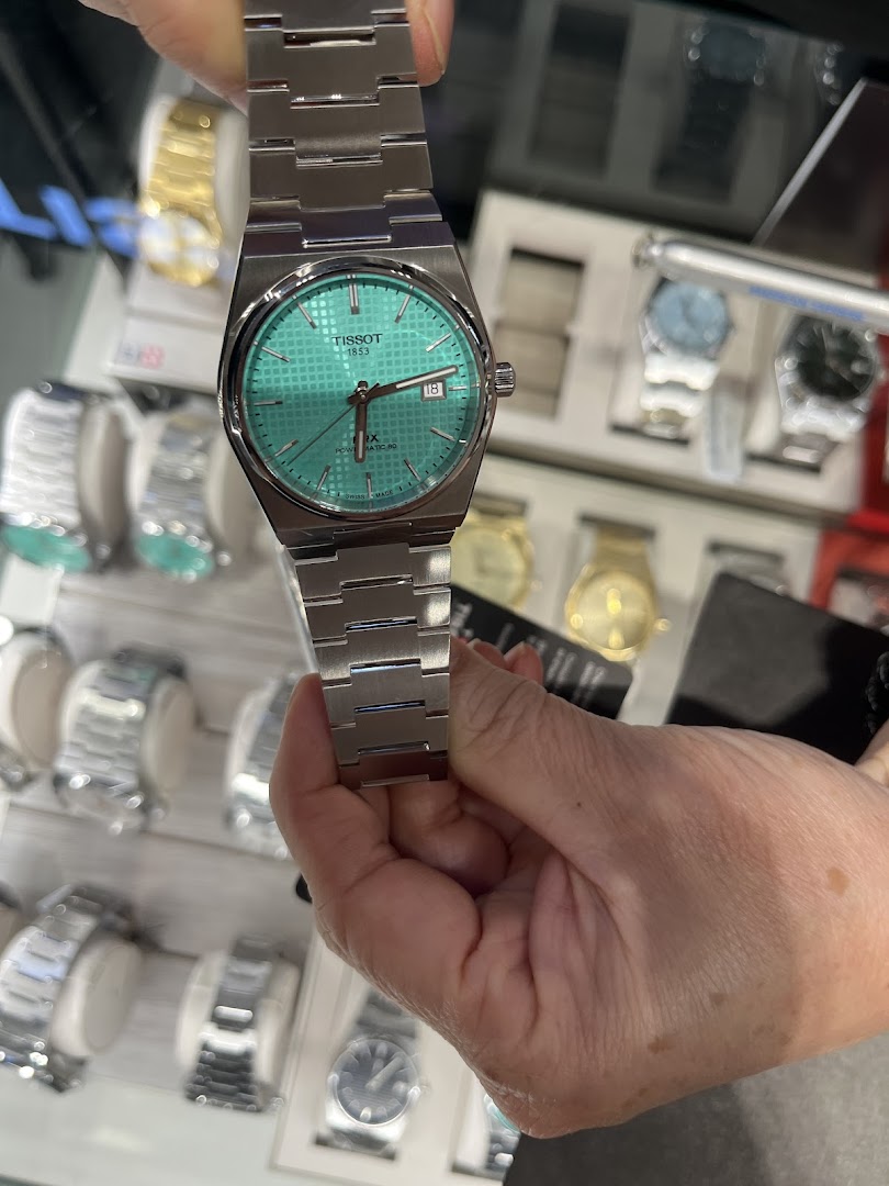 A hand holds a Tissot PRX watch with a striking turquoise dial, showcasing its design.