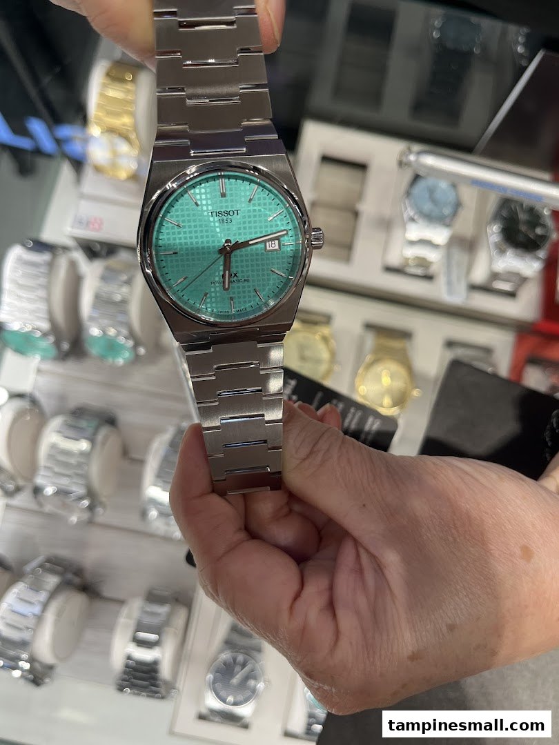 A hand holds a Tissot PRX watch with a striking turquoise dial, showcasing its design.