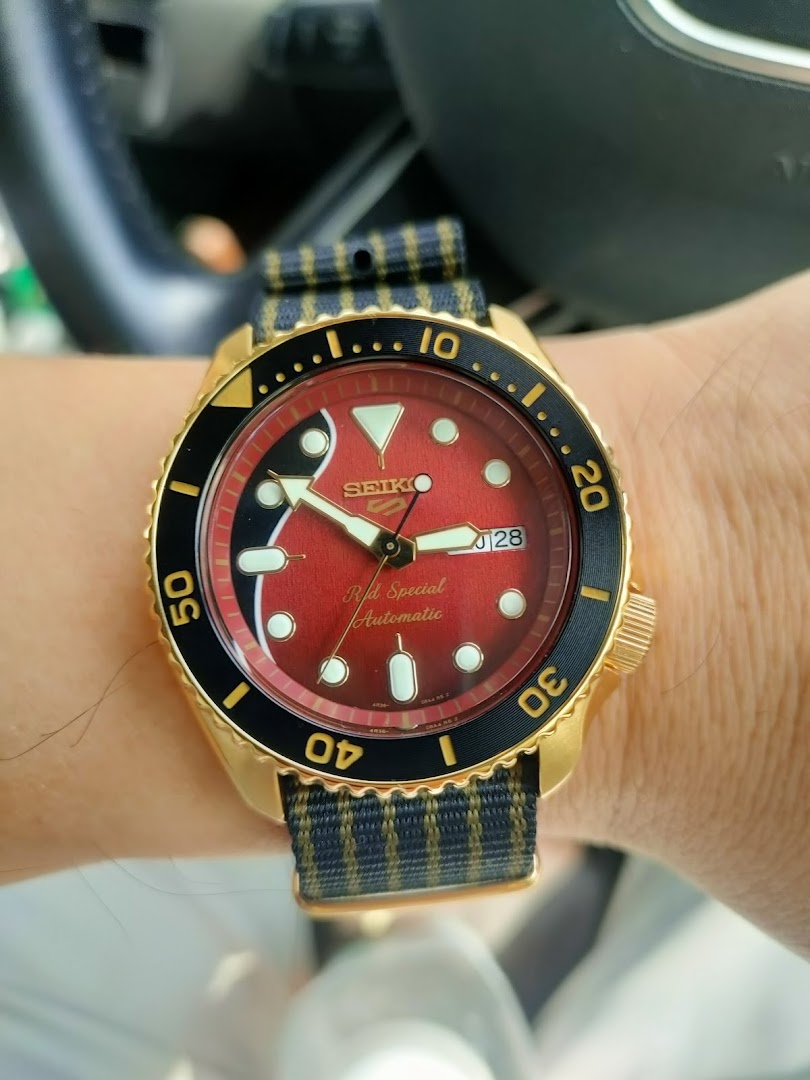 Close-up of a gold-cased Seiko automatic watch with a red dial and a striped fabric strap on a wrist.