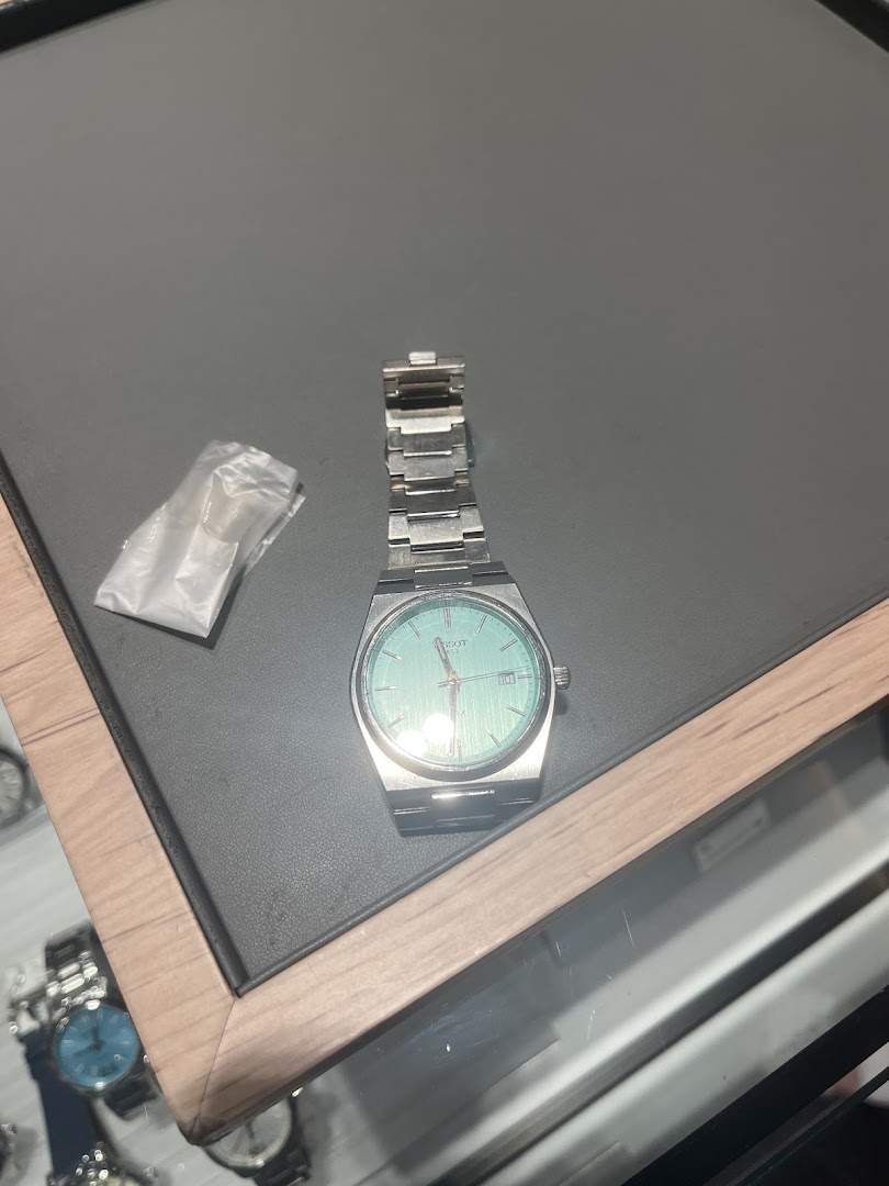 A close-up of a silver watch with a light blue dial displayed on a dark surface, likely in a watch shop.