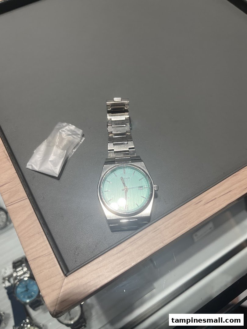 A close-up of a silver watch with a light blue dial displayed on a dark surface, likely in a watch shop.