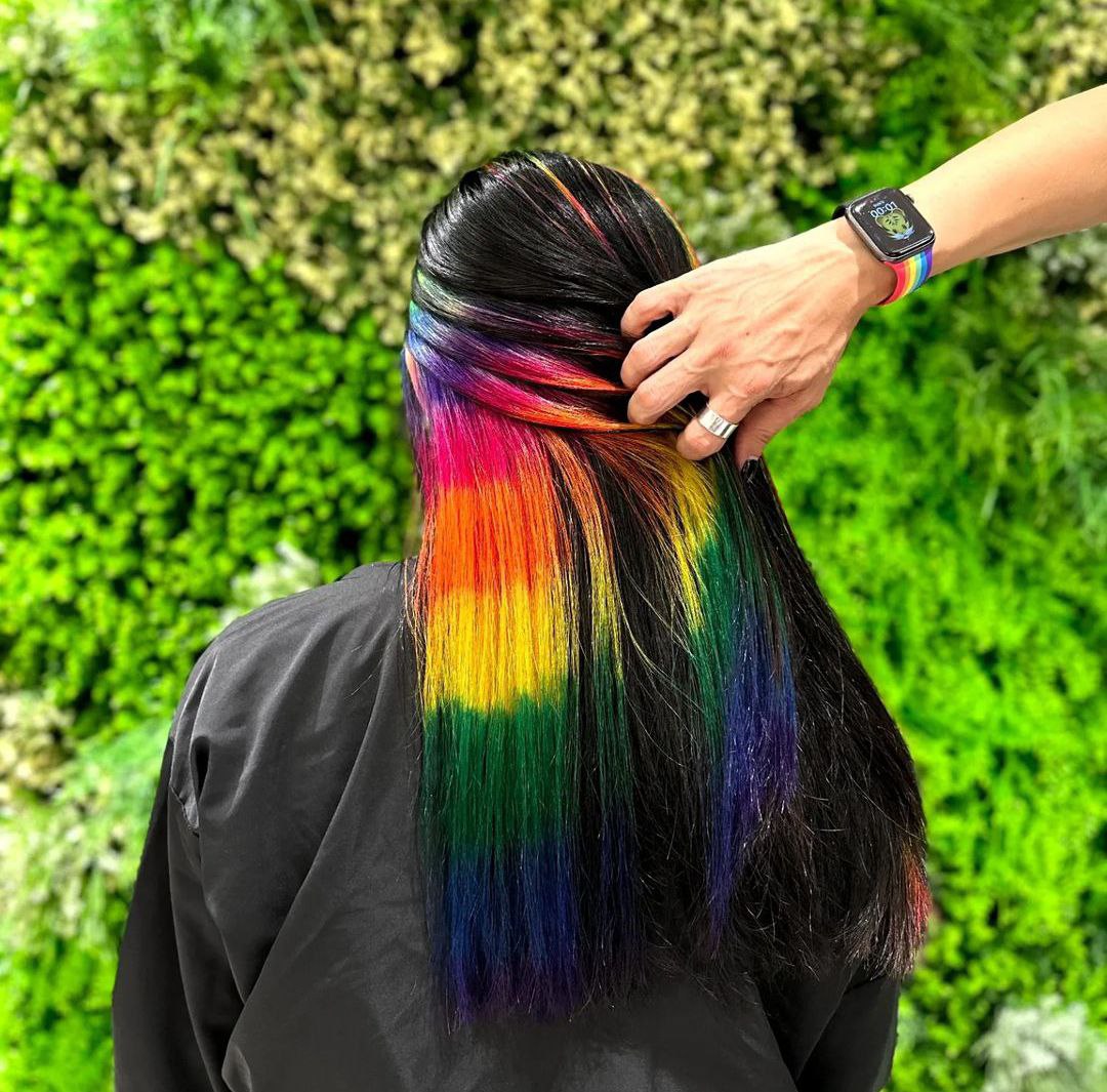 A person's long black hair with vibrant rainbow-colored highlights is being styled against a green wall.