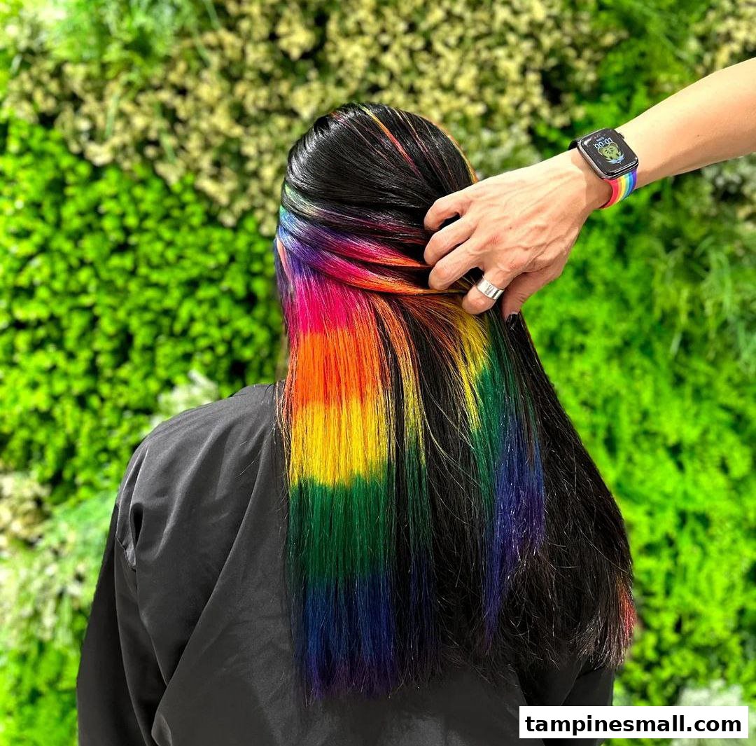 A person's long black hair with vibrant rainbow-colored highlights is being styled against a green wall.