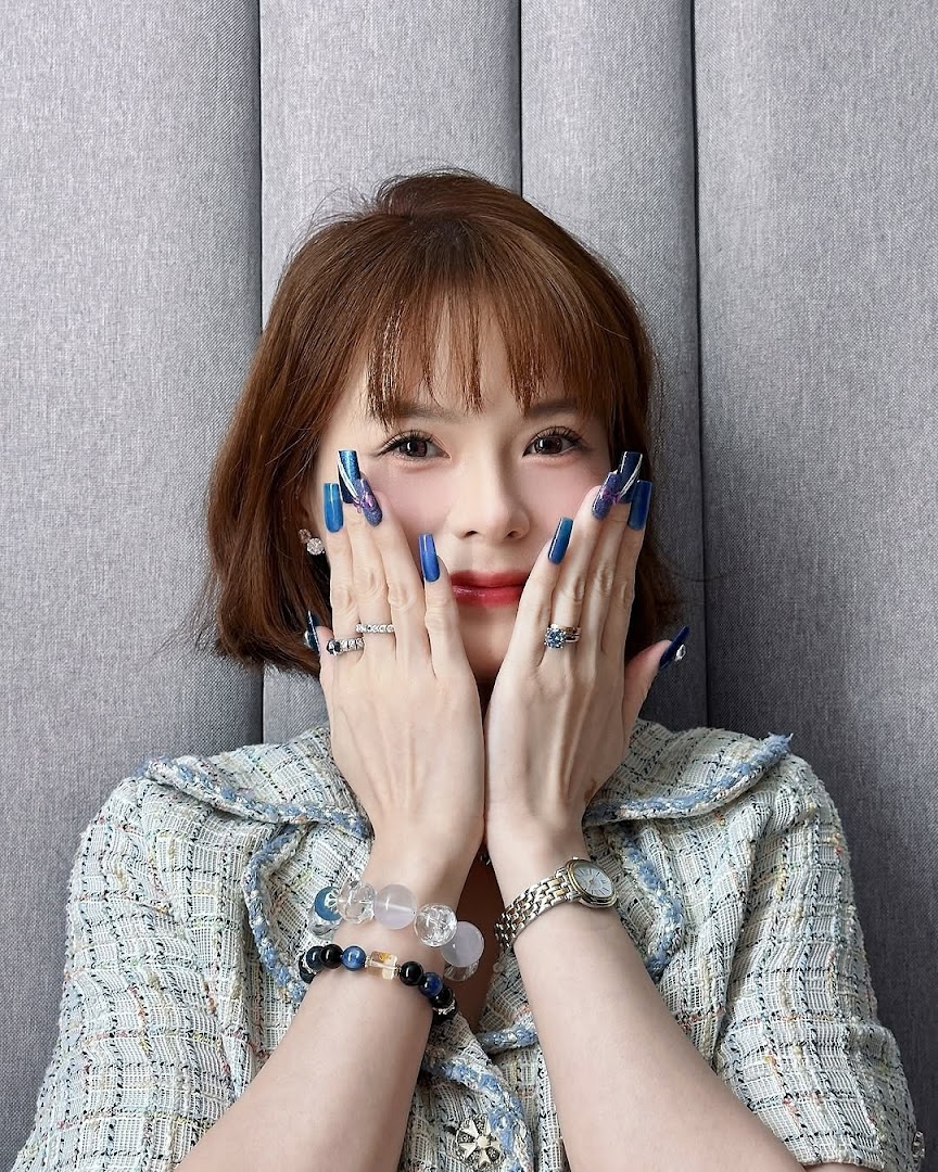 A woman showcasing long, blue manicured nails with intricate designs, wearing rings and bracelets.